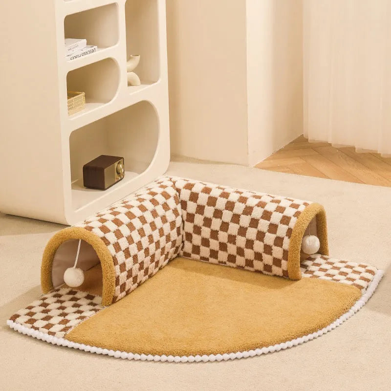 Brown plush plaid cat tunnel bed with central mat on a wooden floor.