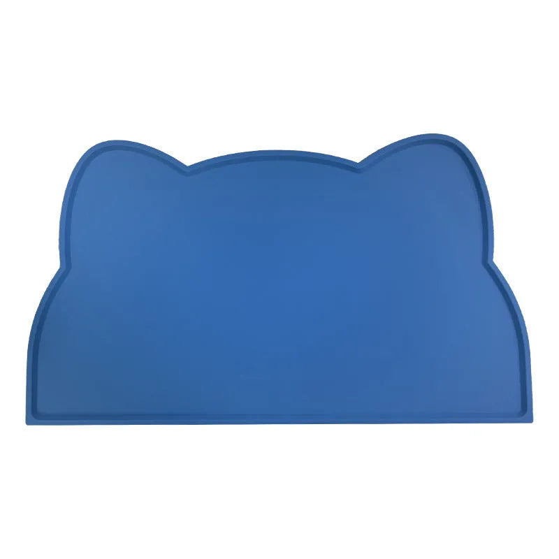 Solid colour silicone pet feeding mat in a cat head shape on a floor next to pet bowls.