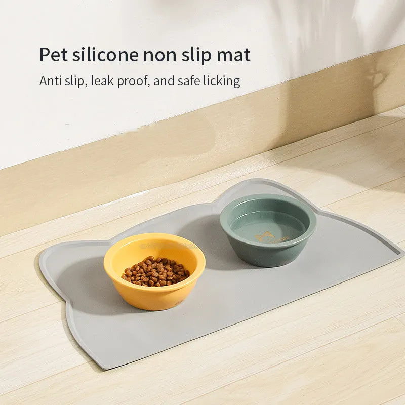 Solid colour silicone pet feeding mat in a cat head shape on a floor next to pet bowls.