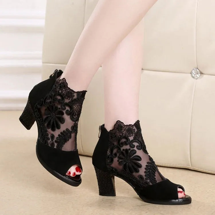 Black lace gladiator sandals with a flower motif, square high heel, and a front zip closure on a woman's foot.