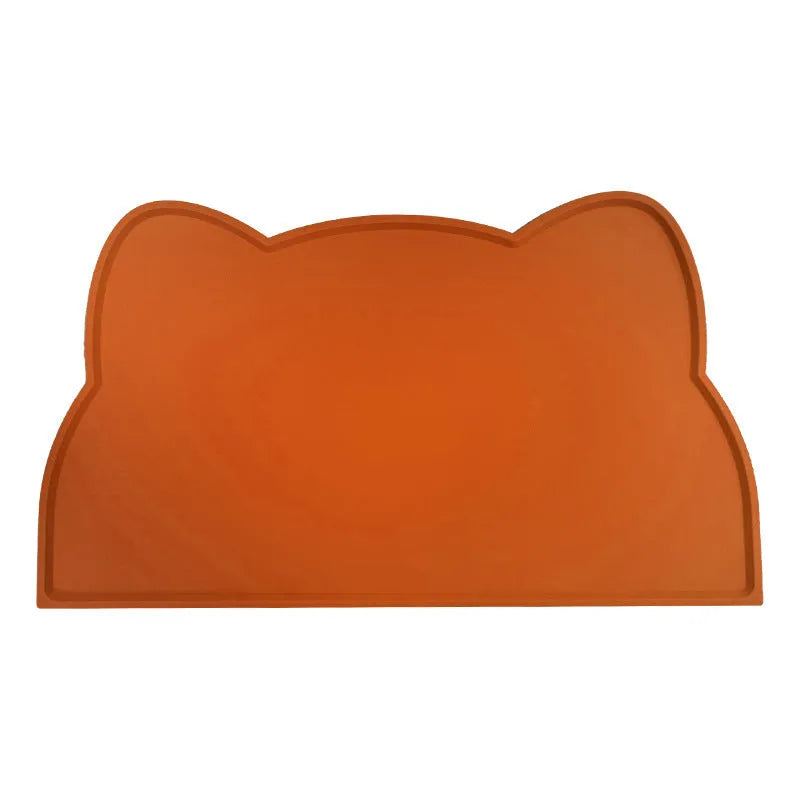 Solid colour silicone pet feeding mat in a cat head shape on a floor next to pet bowls.