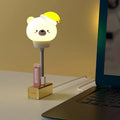 Small cartoon bear-shaped USB night light with remote control on a bedside table.