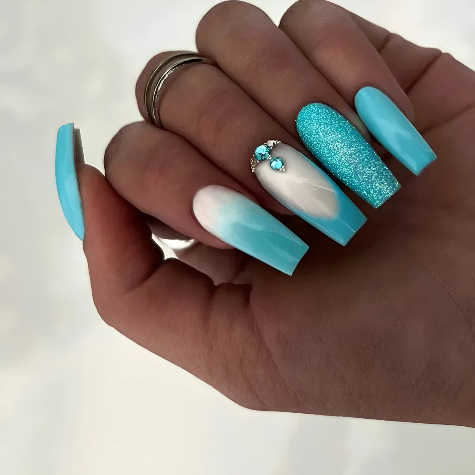 Long ballet shaped artificial nail patches with blue diamond glitter on a solid colour background, shown with a bottle of nail glue.