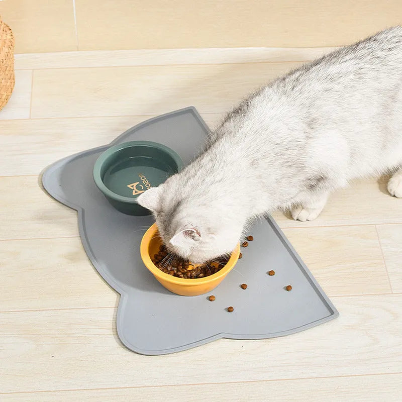 Solid colour silicone pet feeding mat in a cat head shape on a floor next to pet bowls.